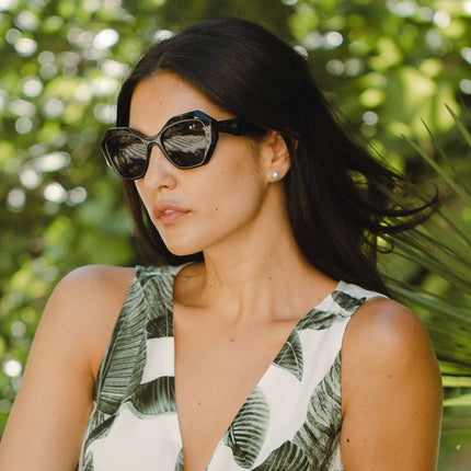 Mujer usando gafas de sol Hanukeii® Moorea Black / Black con fondo de hojas verdes.