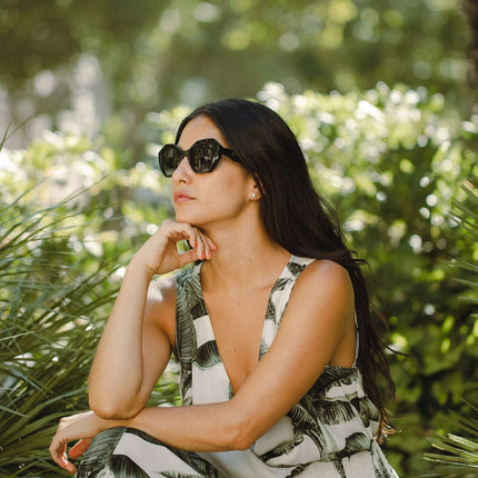 Mujer con gafas de sol Hanukeii® Moorea Black / Black, sentada en un entorno natural con plantas verdes.